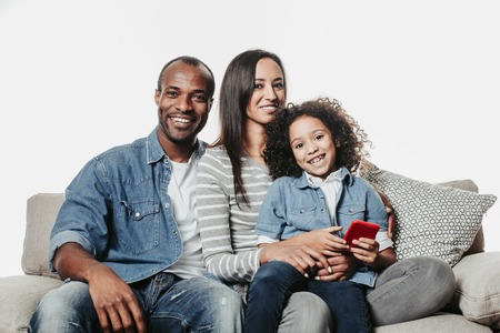 Portrait of laughing mom, daddy and child with device in hand spending free time with comfort. They are looking at camera. Isolated on backgroundの写真素材