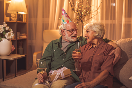 Glad bearded old man embracing happy woman while drinking champagne. They sitting on sofa. Birthday conceptの写真素材