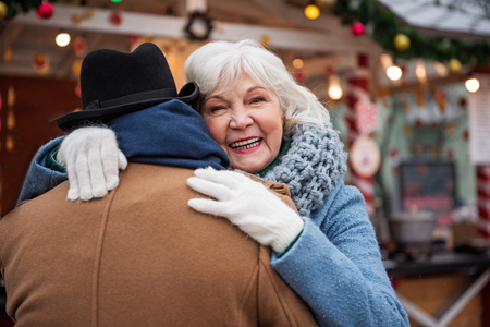 Portrait of happy old woman hugging her husband with love. She is looking at camera and laughing. Focus on male backの写真素材