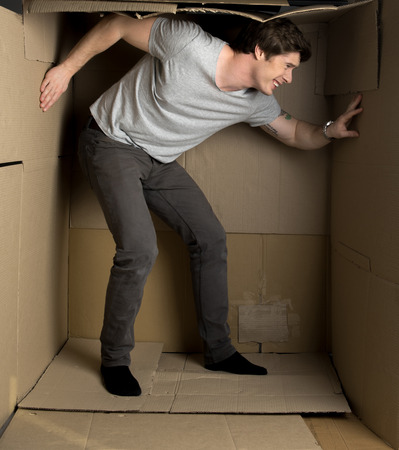 Full length of young irritated man is standing in cramped cardboard box and pressuring on walls while trying to expand them.の写真素材