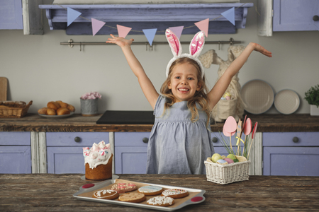 Waist up portrait of happy little girl standing at table with homemade pastry and easter basket, she is raising her arms up and smiling. Country style kitchen on background. Copy space in left sideの写真素材