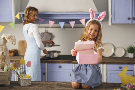 Portrait of glad girl sitting on kitchen table and opening box with easter gift. Mother on background. Focus on kidの写真素材