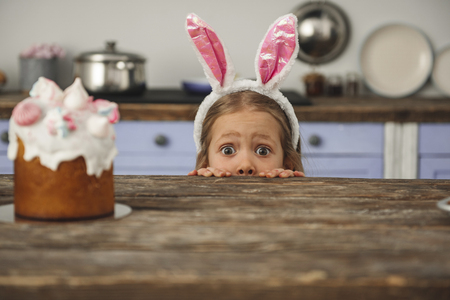 Curious small girl in bunny ears peeking out from behind kitchen table with tasty easter cake and...