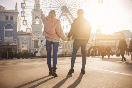 Look here. Full length back view of young man and woman are holding hands while standing on square. Girl is pointing on something. Ferris wheel and sunshine on backgroundの写真素材