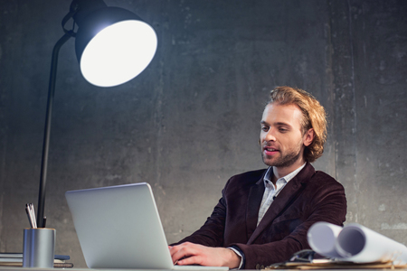 Portrait of outgoing bearded man using laptop while locating at desk during job. Labor with digital device conceptの写真素材