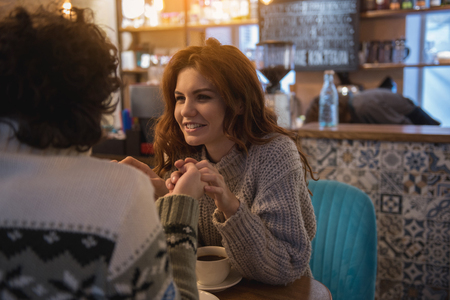 Portrait of glad young red-haired girl looking at her boyfriend with love and smiling. They are touching hands and smiling while sitting in cafeの写真素材