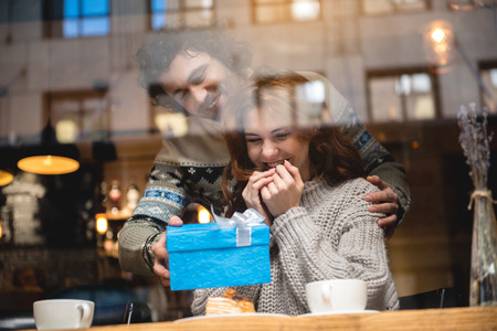 This is for you. Cheerful man is giving present to his wife in cafeteria. Woman is looking at blue...