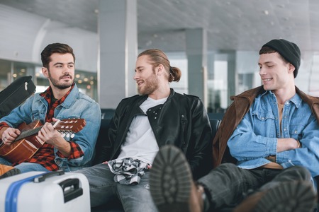 Happy comrades sitting in airport. Beaming man playing guitar while sitting near glad comrades....