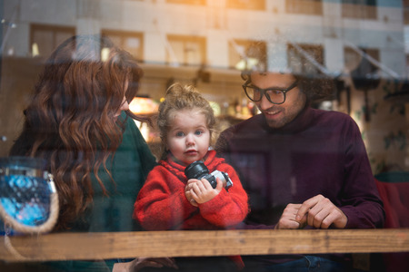 Shocked little girl is holding photo camera in hands. Her parents are looking with her and laughing. Family entertainment conceptの写真素材
