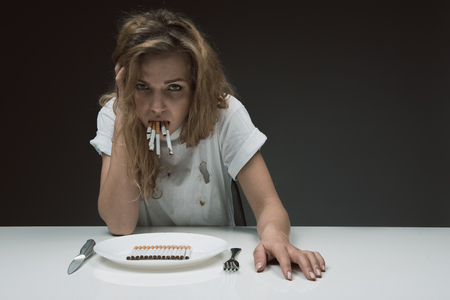 Portrait of displeased lady looking at camera with disgusting face while holding bunch of cigarettes in mouth. Plate with coffin nails on the table. Copy space in right side. Isolated on backgroundの写真素材