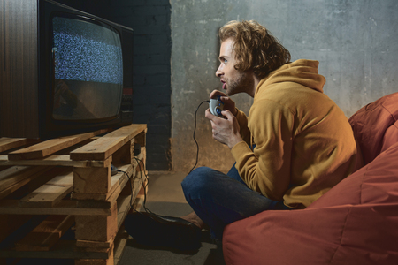 Side view excited bearded man playing in video game while sitting on armchair in room. Entertainment conceptの写真素材