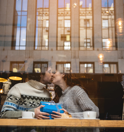 Happy Valentine day. Cheerful loving couple kissing in cafeteria. They are holding big present box while sitting near the windowの写真素材