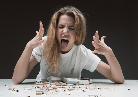 Portrait of impetuous woman sitting at table with smashed cigarettes, she is holding hands up and making wry face in disgust. Isolated on backgroundの写真素材