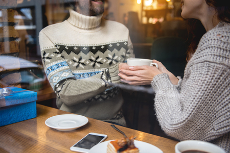 Happy loving couple holding a cup of coffee in hands together. They are sitting in cafe and smiling. Gift box is on tableの写真素材