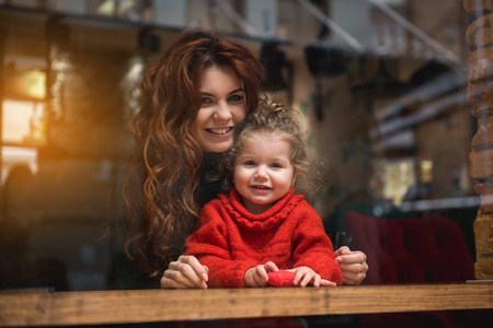 Portrait of happy mother embracing her little daughter while sitting at table. They are looking forward over the window and smilingの写真素材