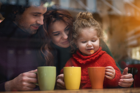 Portrait of happy girl looking inside cup of tea with interest. Her mother and father are embracing the child while sitting in cafe and smilingの写真素材