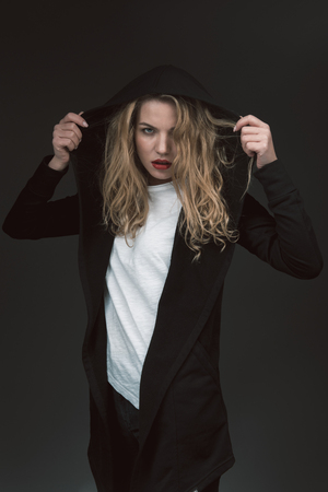 Waist up portrait of confident blonde female in black cardigan putting on hood on her head. Isolated on backgroundの写真素材