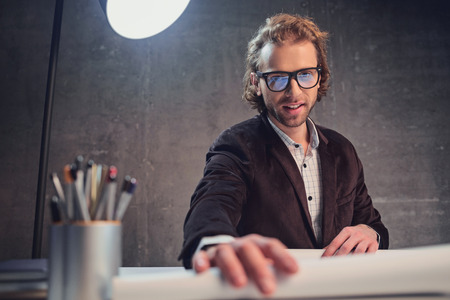 Portrait of happy unshaven male architect sitting at table while looking in camera. Paperwork conceptの写真素材