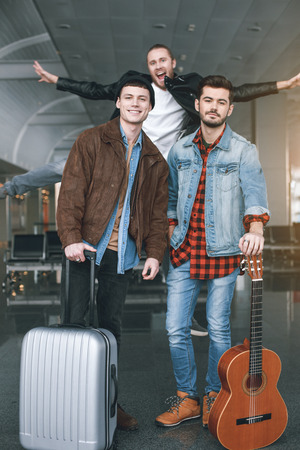 Portrait of smiling young friends with baggages and guitar situating in hall. Outgoing male jumping there. Journey conceptの写真素材