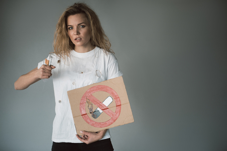 Waist up portrait of serious young woman clenching tobacco in fist and holding no smoking sign on paper. Copy space in right side. Isolated on backgroundの写真素材