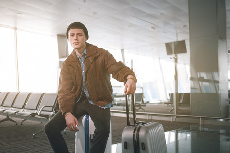 Portrait of weariful young man sitting on luggage while looking in camera in airport. Anticipation conceptの写真素材