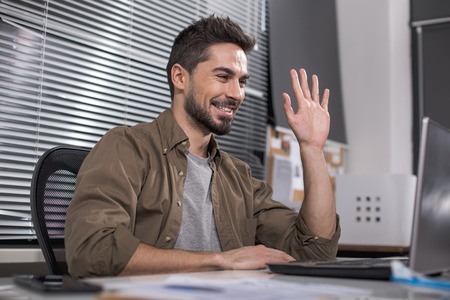 Joyful handsome male in office looking at laptop screen and waving his hand cheerfully
