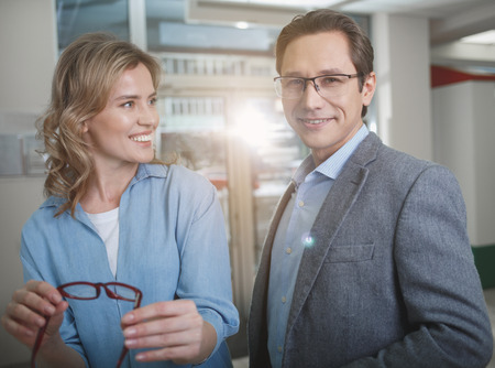 Portrait of happy woman and outgoing male situating in optician store. Female holding eyeglasses in arms. Eyesight correction conceptの写真素材