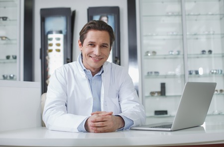 Portrait of glad young doctor sitting at table with notebook computer indoor. Medicine conceptの写真素材