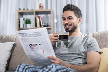 Portrait of contented handsome male reading news on couch and holding cup of coffeeの写真素材