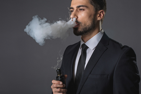 Waist up portrait of pensive young man electronic cigarette. He is standing and looking far away. Isolated on grey backgroundの写真素材