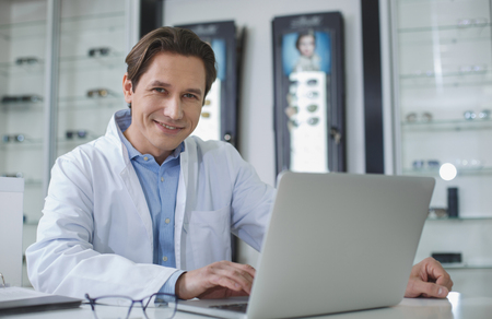 Portrait of beaming male ophthalmologist working with laptop while looking at camera. Optician store conceptの写真素材