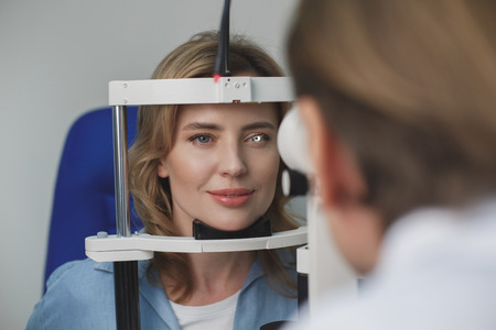 Portrait of smiling female client checking eyesight with equipment opposite doctor. Ophthalmology conceptの写真素材