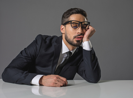 Waist up of dozing exhausted young man sitting at the white desk with head reclined upon handの写真素材