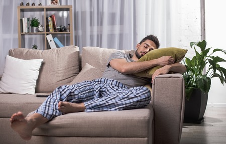 Tranquil young male sleeping on comfortable couch in living room. Bookshelf on backgroundの写真素材