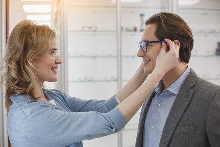 Side view beaming female wearing eyeglasses on male. They looking at each other. Glad couple in optician store conceptの写真素材