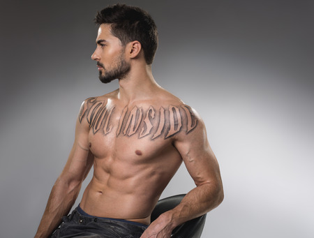 Masculinity concept. Profile of serious young man with strong body sitting on chair without shirt. Isolated on backgroundの写真素材