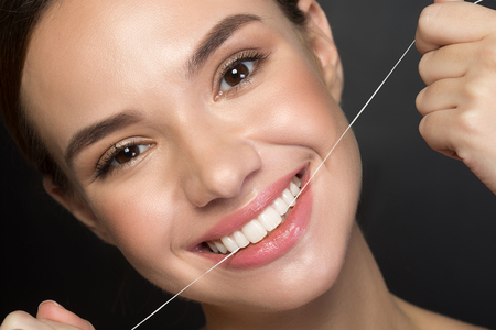Close up portrait of cheerful girl is flossing her teeth by special thread. She is looking at camera...