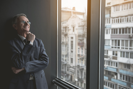 Portrait of pensive senior employer looking at window while leaning against wall indoor. He keeping chin by hand. Dreaminess during work conceptの写真素材
