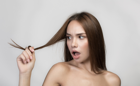 Disappointed young woman with naked shoulders is standing and holding damaged ends of her hair in hand. Isolated background. Haircare conceptの写真素材