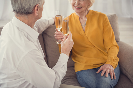 Memorable date. Satisfied old couple sitting with comfort at home and holding glasses full of champagneの写真素材