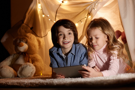 Joyous children using modern tablet in playroom. They lying in toy tent on carpet. Boy looking at camera with happinessの写真素材