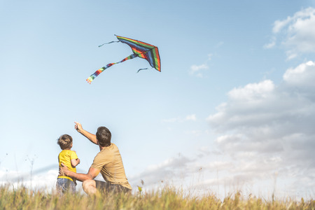 Parent and little boy are sitting with their backs to camera on field and flying kite. Copy space in right sideの写真素材