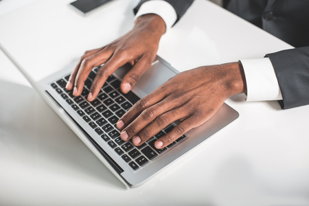 Important project. Top view close up of hands of young african entrepreneur is sitting at his workplace and typing on modern laptop. Selective focusの写真素材