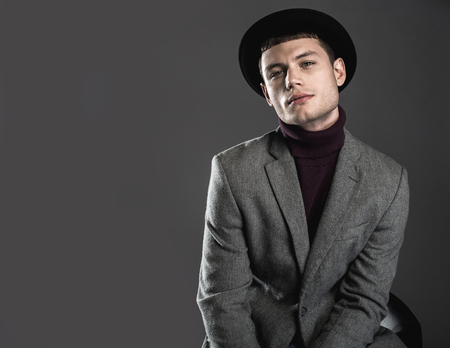 Portrait of concentrated man wearing contemporary hat while sitting on chair. He isolated on black background. Copy space. Focused fashionable businessman conceptの写真素材
