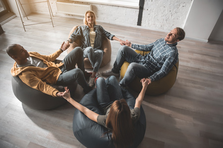 Top view of serene men and women sitting in circle on beanbags with eyes closed. They are holding...
