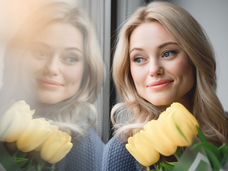 Portrait of dreamful young woman looking over the window while holding yellow tulips