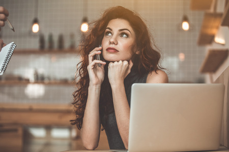 Portrait of pensive young woman is talking on phone while looking at waiter in cafe. She is sitting near laptop in cafeteriaの写真素材