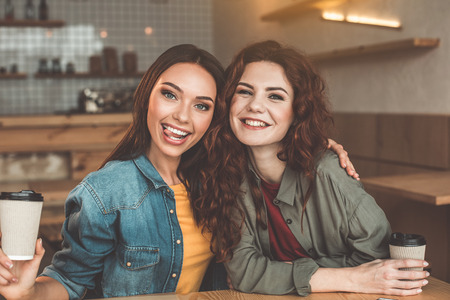 Best friends. Waist up portrait of cheerful young girls drinking coffee in cafe. They are embracing and smilingの写真素材
