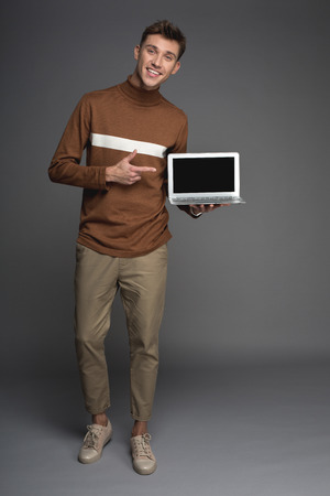 Full length portrait of smiling male person standing and pointing at laptop in his armの写真素材