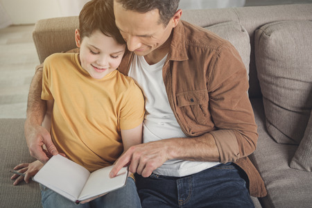 This is my favorite moment. Top view of caring father pointing finger at book and embracing son. Boy...